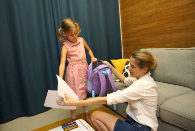 Genç anne, kızının okul için sırt çantasını toplamasına yardım ediyor.