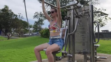 Genç bir kadın, doğa manzaralı bir parkta, modern egzersiz aletleriyle dışarıda çalışmaktan zevk alıyor.