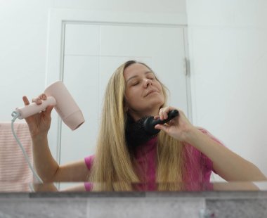 Bir kadın uzun saçlarını kuruturken aynı zamanda onu tarıyor.