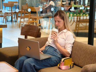 Genç bir kadın canlı bir kafede dizüstü bilgisayarıyla çalışırken kahveden zevk alır.