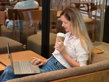 Genç bir kadın canlı bir kafede dizüstü bilgisayarıyla çalışırken kahveden zevk alır.