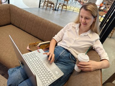 Genç bir kadın canlı bir kafede dizüstü bilgisayarıyla çalışırken kahveden zevk alır.