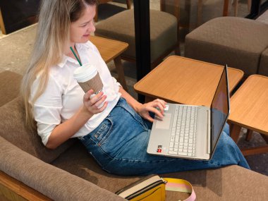 Genç bir kadın canlı bir kafede dizüstü bilgisayarıyla çalışırken kahveden zevk alır.