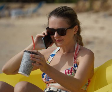 Kumsalda genç bir kadın, elinde bir içki ve akıllı telefonuyla sarı bir fasulye torbasının üzerinde uzanmış. 