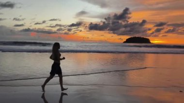 Gün batımında sakin sahil sahnesinde koşan ve yürüyen bir kadın. 