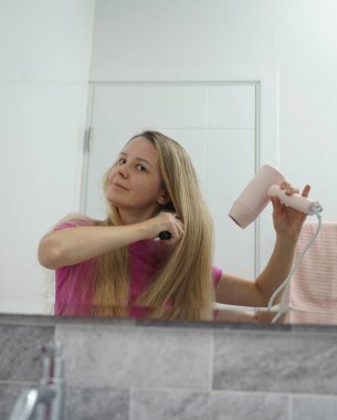 Bir kadın uzun saçlarını kuruturken aynı zamanda onu tarıyor.