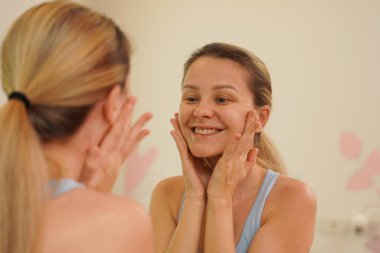 Modern bir banyoda genç bir kadın yüzünü yıkıyor ve sabahları dişlerini fırçalıyor.