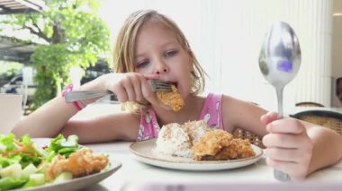 Neşeli küçük bir kız kafede kızarmış tavuk ve taze salata yiyor.