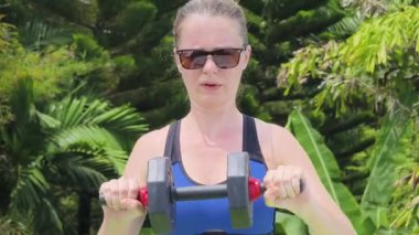  Genç bir kadın yemyeşil bir ortamda yoga minderinin üzerinde halterlerle antrenman yapıyor.