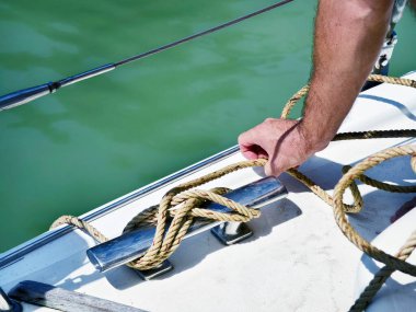 Yelkenlinin güvertesinde erkek kaptan yelkenleri açıyor halatı çekiyor.