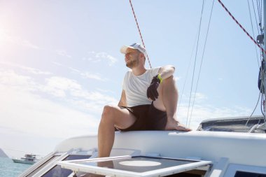 Bir adam rahat bir şekilde bir yelkenlinin güvertesinde oturmuş hafif esintinin tadını çıkarıyor.