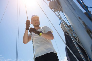 Yelkenlinin güvertesinde erkek kaptan yelkenleri açıyor halatı çekiyor.