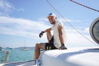 Bir adam rahat bir şekilde bir yelkenlinin güvertesinde oturmuş hafif esintinin tadını çıkarıyor.