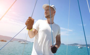 Yelkenlinin güvertesinde erkek kaptan yelkenleri açıyor halatı çekiyor.