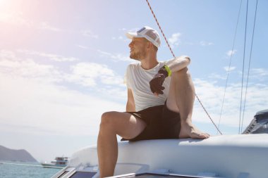 Bir adam rahat bir şekilde bir yelkenlinin güvertesinde oturmuş hafif esintinin tadını çıkarıyor.