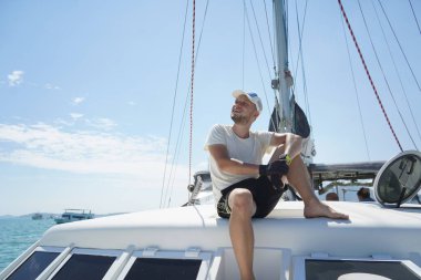 Bir adam rahat bir şekilde bir yelkenlinin güvertesinde oturmuş hafif esintinin tadını çıkarıyor.