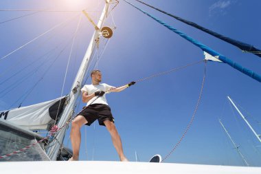 Yelkenlinin güvertesinde erkek kaptan yelkenleri açıyor halatı çekiyor.