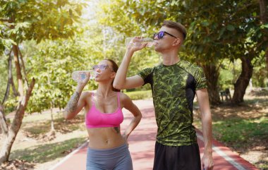 Bir adam ve bir kadın parkta birlikte koştuktan sonra şişelerden ferahlatıcı su içiyorlar..