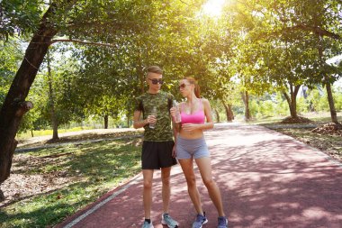 Bir adam ve bir kadın parkta birlikte koştuktan sonra şişelerden ferahlatıcı su içiyorlar..