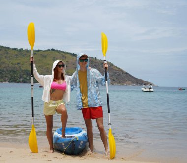 Kumsalda genç bir çift ellerinde kano kürekleri ve suda eğlenceli bir yolculuğa hazırlar..