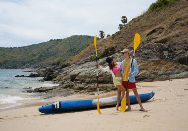 Kumsalda genç bir çift ellerinde kano kürekleri ve suda eğlenceli bir yolculuğa hazırlar..