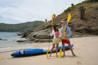 Kumsalda genç bir çift ellerinde kano kürekleri ve suda eğlenceli bir yolculuğa hazırlar..