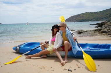 Kumsalda genç bir çift ellerinde kano kürekleri ve suda eğlenceli bir yolculuğa hazırlar..