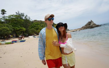 Genç bir çift kumsalda boş boş yürüyor, el ele tutuşuyor ve sıcak güneşin tadını çıkarıyor..