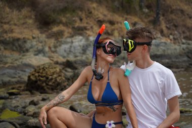 Genç bir çift, su kenarındaki bir kayanın üzerinde otururken gözlük ve şnorkel takıyor..