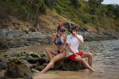 Genç bir çift, su kenarındaki bir kayanın üzerinde otururken gözlük ve şnorkel takıyor..