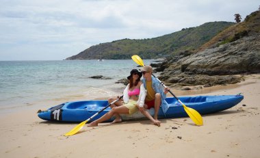 Kumsalda genç bir çift ellerinde kano kürekleri ve suda eğlenceli bir yolculuğa hazırlar..