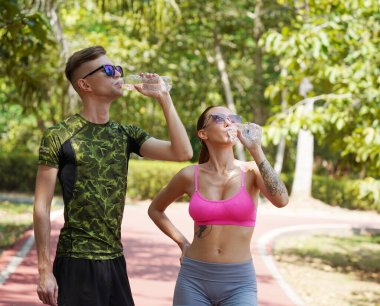 Bir adam ve bir kadın parkta birlikte koştuktan sonra şişelerden ferahlatıcı su içiyorlar..