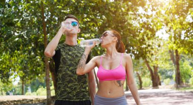 Bir adam ve bir kadın parkta birlikte koştuktan sonra şişelerden ferahlatıcı su içiyorlar..