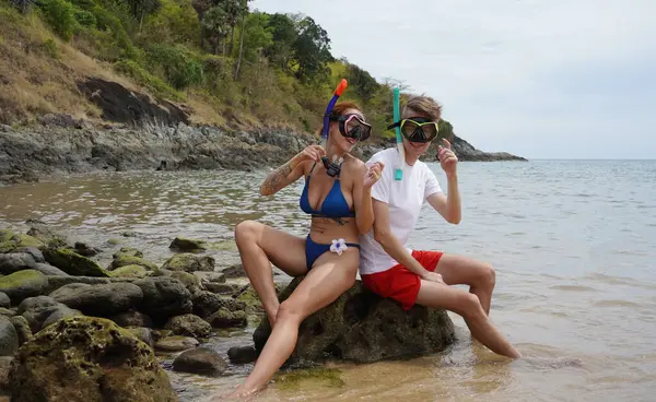 Genç bir çift, su kenarındaki bir kayanın üzerinde otururken gözlük ve şnorkel takıyor..