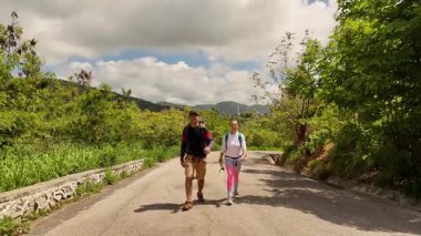 Bir erkek ve bir kadın, doğa güzelliği ve sükunetle çevrili bir yolda rahatça yürüyorlar..