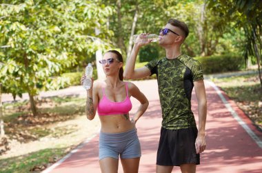 Bir adam ve bir kadın parkta birlikte koştuktan sonra şişelerden ferahlatıcı su içiyorlar..