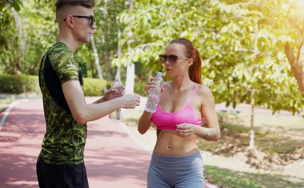 Bir adam ve bir kadın parkta birlikte koştuktan sonra şişelerden ferahlatıcı su içiyorlar..