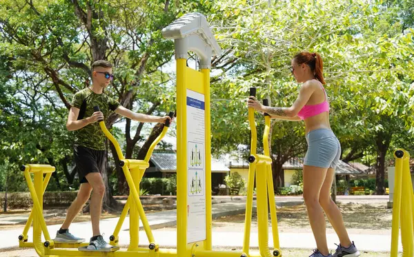 Bir adam ve kadın parkta bir makinede birlikte egzersiz yapıyorlar..