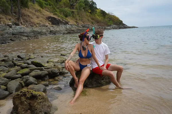 Genç bir çift, su kenarındaki bir kayanın üzerinde otururken gözlük ve şnorkel takıyor..