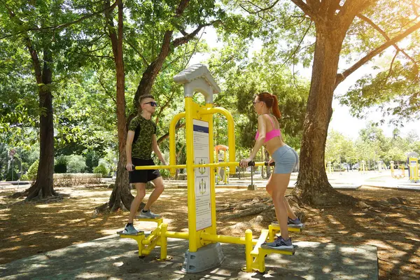 Bir adam ve kadın parkta bir makinede birlikte egzersiz yapıyorlar..