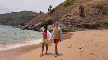 Genç bir çift kumsalda boş boş yürüyor, el ele tutuşuyor ve sıcak güneşin tadını çıkarıyor..