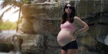 Hamile bir kadın zarif bir şekilde güzel bir şelale kayasının önünde duruyor..