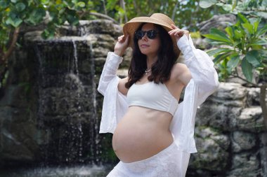 Hamile bir kadın zarif bir şekilde güzel bir şelale kayasının önünde duruyor..