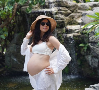 Hamile bir kadın zarif bir şekilde güzel bir şelale kayasının önünde duruyor..