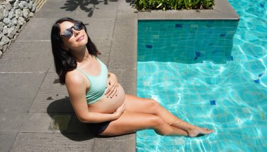 Turkuaz bikinili hamile bir kadın havuz kenarında oturuyor..
