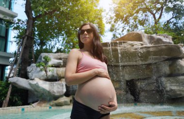 Hamile bir kadın zarif bir şekilde güzel bir şelale kayasının önünde duruyor..