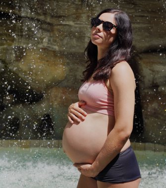 Hamile bir kadın zarif bir şekilde güzel bir şelale kayasının önünde duruyor..