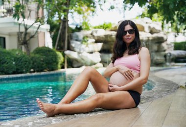 Pembe bikinili hamile bir kadın havuzun kenarında oturuyor..