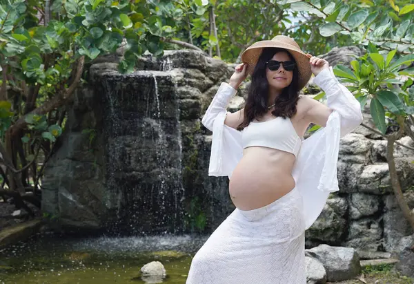 Hamile bir kadın zarif bir şekilde güzel bir şelale kayasının önünde duruyor..