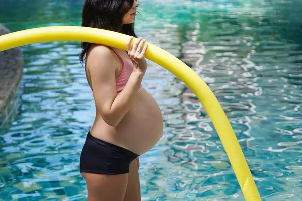 Bilardo oynamak üzere olan hamile genç kadın köpüklü makarnayla havuzda egzersiz yapacak..